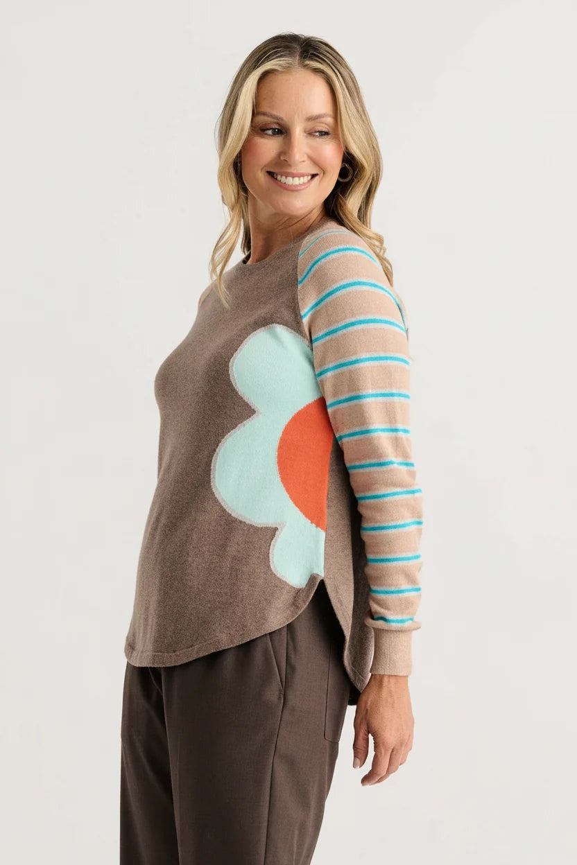 A woman modeling a brown knit sweater with a large light blue and orange flower applique on the front and one striped sleeve in beige, brown, and turquoise blue.