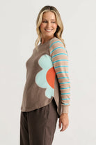 A woman modeling a brown knit sweater with a large light blue and orange flower applique on the front and one striped sleeve in beige, brown, and turquoise blue.