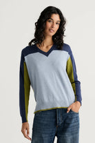 A woman wearing a color-blocked knit sweater with a light blue body, navy blue shoulders and sleeves, and olive green side panels, paired with blue jeans against a plain light background.
