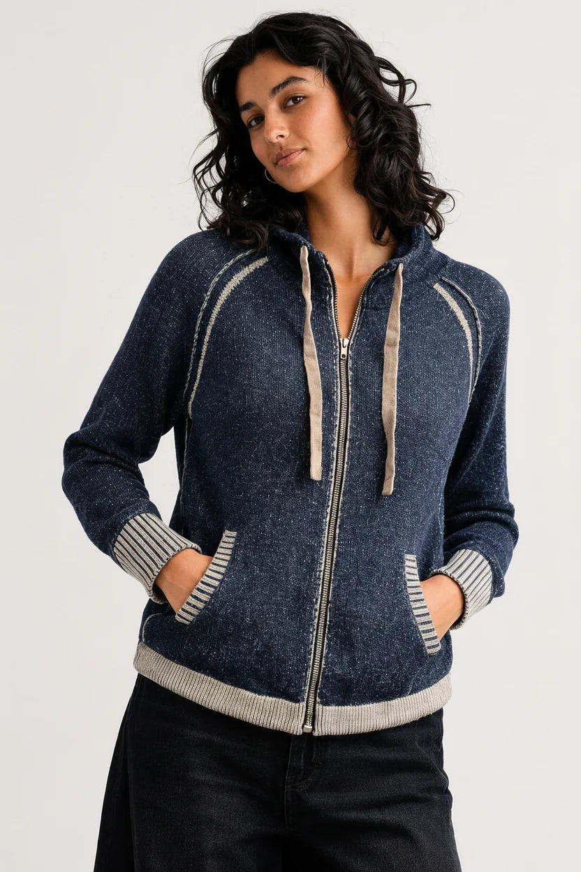 A woman wearing a dark blue zip-up hoodie with beige ribbed trim and drawstrings, paired with black pants, photographed against a light gray studio background.