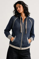 A woman wearing a dark blue zip-up hoodie with beige ribbed trim and drawstrings, paired with black pants, photographed against a light gray studio background.