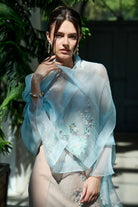 A woman wearing a sheer light blue embroidered shawl draped over her shoulders, paired with a beige dress, set against a background of green foliage and a dark wooden door.