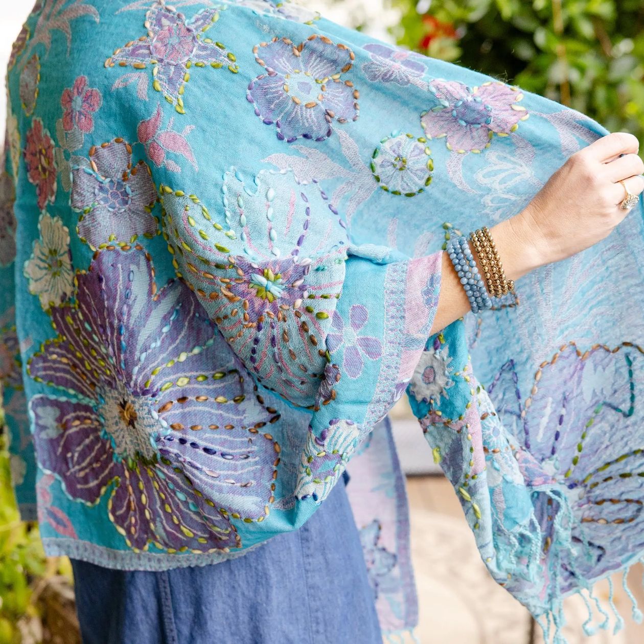 A light blue bohemian shawl with large purple and lavender floral embroidery, held up by a model wearing blue jeans and beaded bracelets.