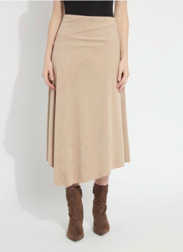A woman modeling a caramel vegan suede midi skirt with a fitted waist, diagonal seaming, and a raw-edge asymmetric hem, paired with a black top and brown suede boots, against a plain light studio background.
