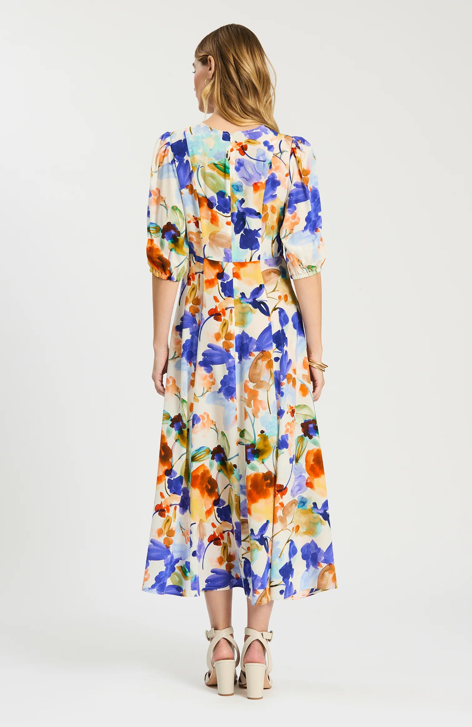 A woman viewed from the back wearing a midi dress with a colorful watercolor floral print in blue, orange, yellow, and green on a white base, paired with beige strappy high-heeled sandals against a light gray studio background.