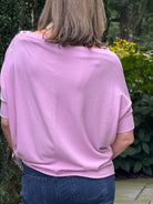 A woman viewed from behind wearing a light pink short-sleeve top and blue jeans, standing outdoors in a garden setting with green foliage and yellow flowers in the background.