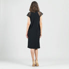 A woman viewed from behind wearing a black knee-length dress with sheer mesh cap sleeves, paired with beige high-heeled sandals, against a light gray studio background.