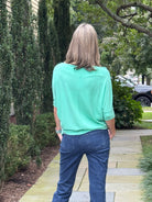 A woman viewed from behind wearing a mint green short-sleeve top and blue jeans, standing on a stone pathway in a garden setting.