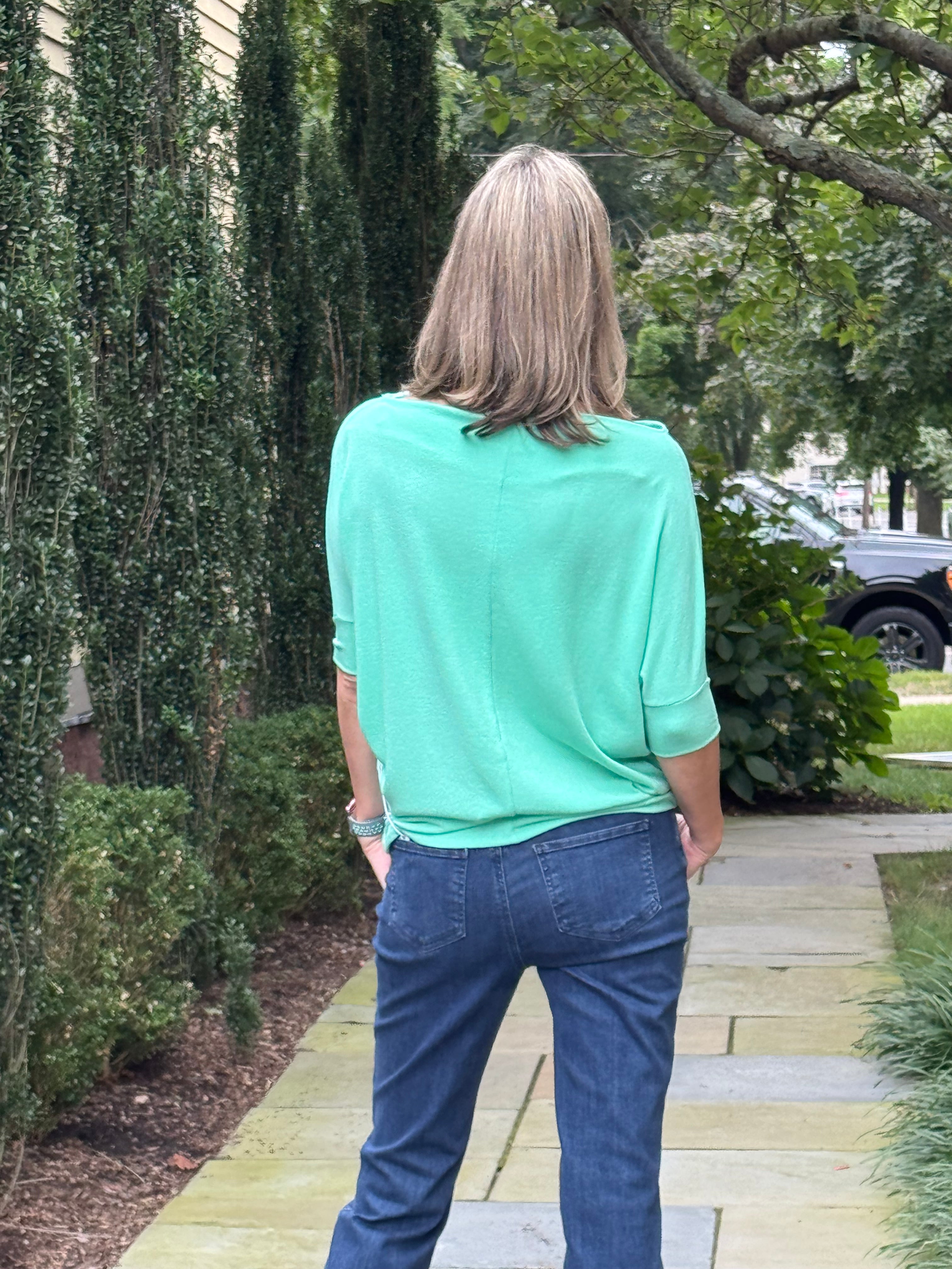 A woman viewed from behind wearing a mint green short-sleeve top and blue jeans, standing on a stone pathway in a garden setting.