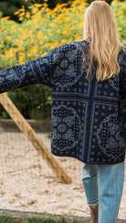 A woman viewed from behind wearing a navy blue bandana print cardigan over a light blue top and light wash jeans, standing outdoors in front of yellow flowers.