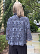 A woman viewed from behind wearing a blue-gray lace top with a high mock neck and three-quarter sleeves, paired with black pants, standing outdoors on a stone walkway.