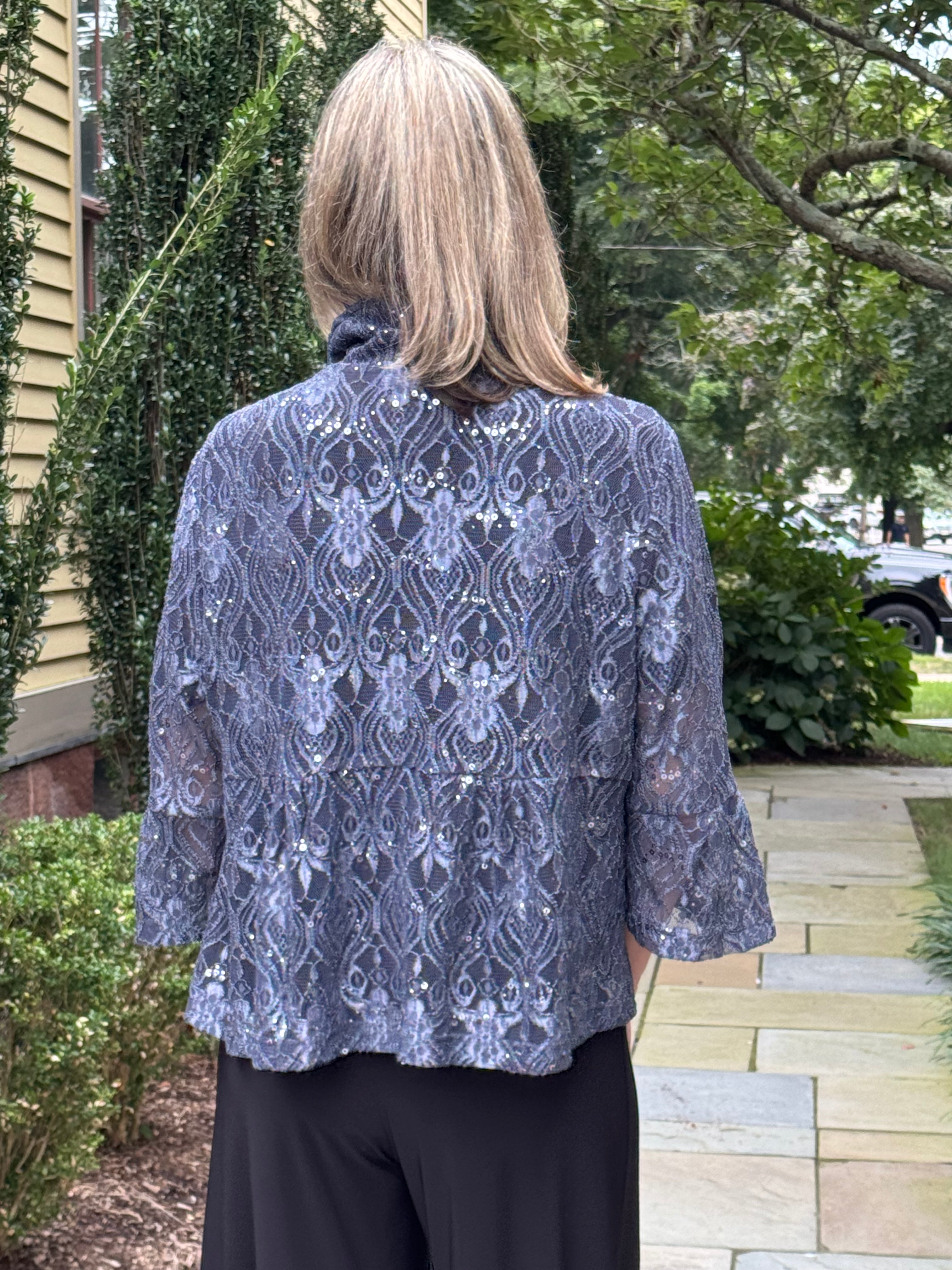 A woman viewed from behind wearing a blue-gray lace top with a high mock neck and three-quarter sleeves, paired with black pants, standing outdoors on a stone walkway.