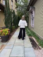 A woman viewed from behind walking on a stone path between houses, wearing a white puff-sleeve blouse and black wide-leg pants.