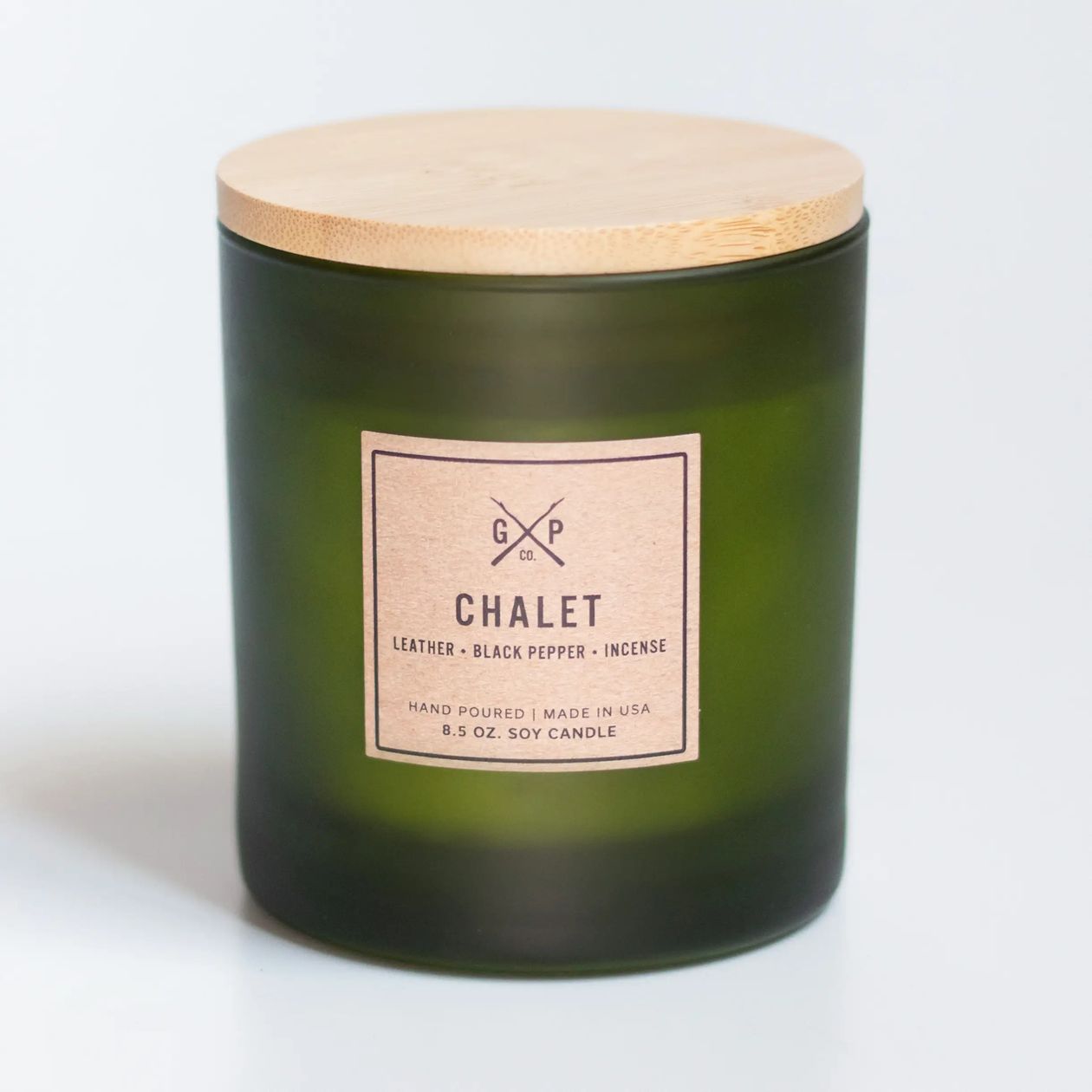 A dark green frosted glass soy candle jar with a light wood lid and a beige paper label reading 'CHALET' and 'GXP CO.', photographed on a clean white background.
