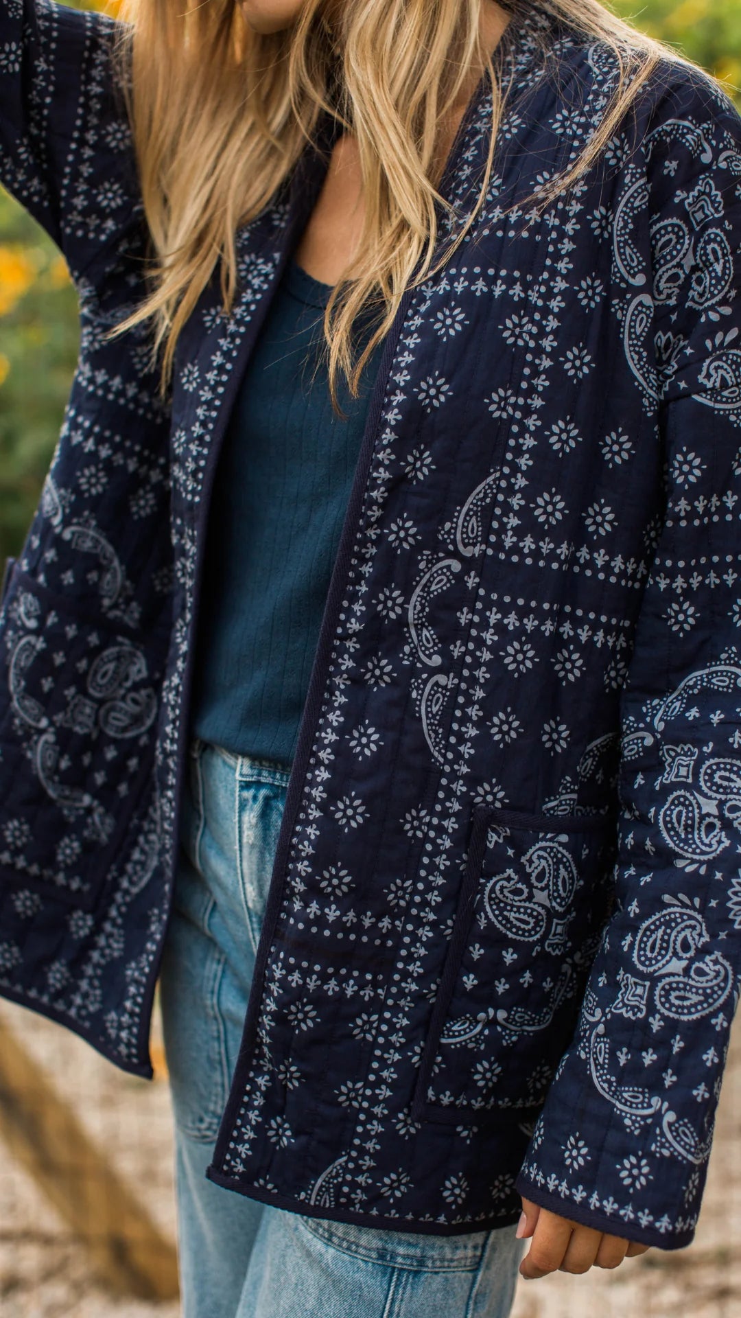 A close-up of a person wearing a navy blue quilted jacket with a white paisley and geometric bandana print, layered over a dark teal top and light blue jeans.