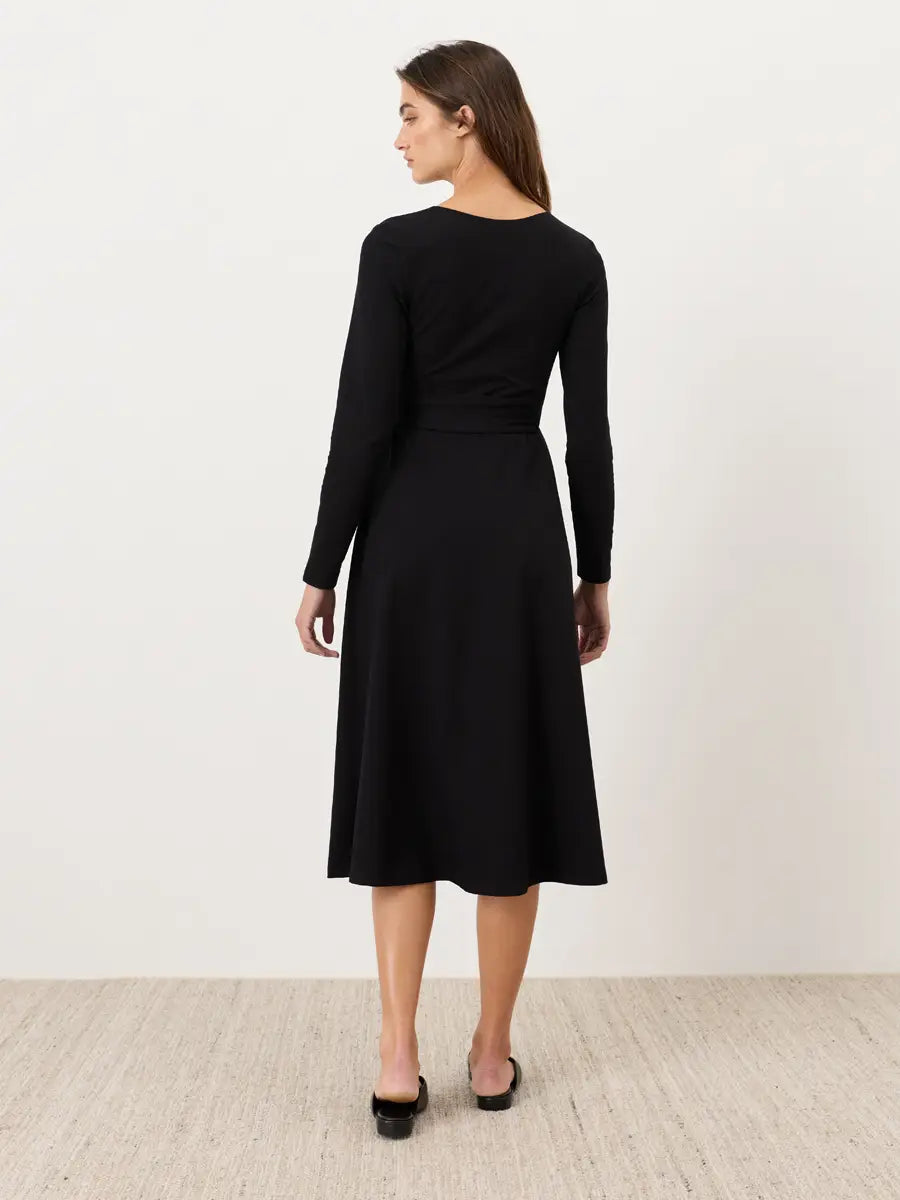 A woman wearing a black long-sleeve midi dress, shown from the back against a plain off-white wall and light wood floor.