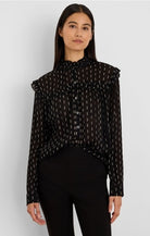 A woman modeling a sheer black long-sleeve blouse with a small white geometric pattern, ruffled shoulders, and a high gathered neckline, paired with black pants against a light gray studio background.