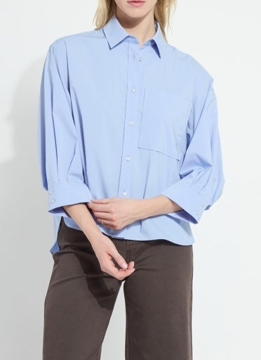 A model wearing a light blue button-down shirt with three-quarter sleeves and dark brown trousers, shown from the neck down against a plain white background.