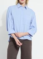 A model wearing a light blue button-down shirt with three-quarter sleeves and dark brown trousers, shown from the neck down against a plain white background.