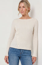 A woman wearing a cream ribbed knit long-sleeve button-front top and blue jeans, standing against a plain light background.