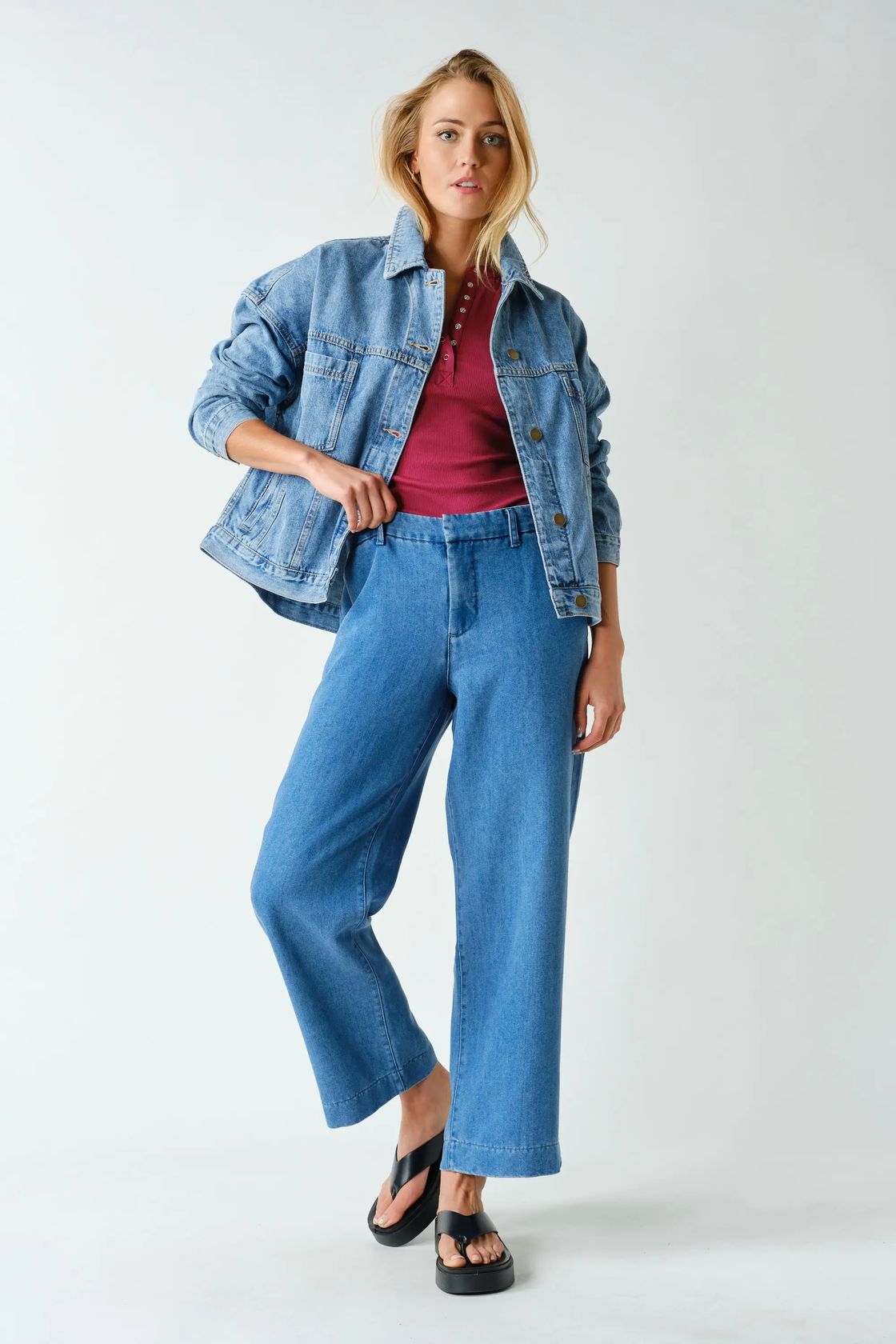 A woman modeling a light blue denim jacket and matching wide-leg cropped jeans over a burgundy top, paired with black sandals, against a plain light gray studio background.