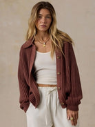 A woman wearing a rust-brown ribbed knit button-up cardigan over a white tank top and white drawstring pants, standing against a textured beige wall.
