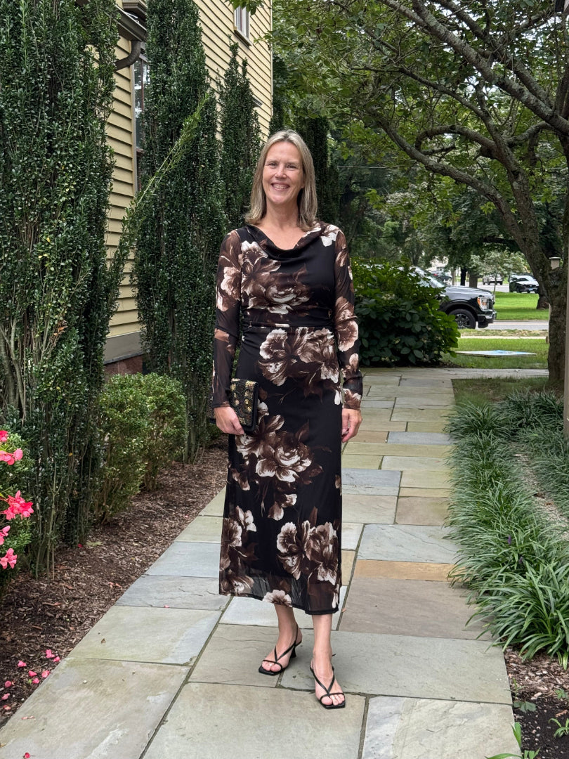 A woman stands on a stone walkway in a landscaped garden, wearing a black midi dress with a brown and beige floral pattern and sheer long sleeves.