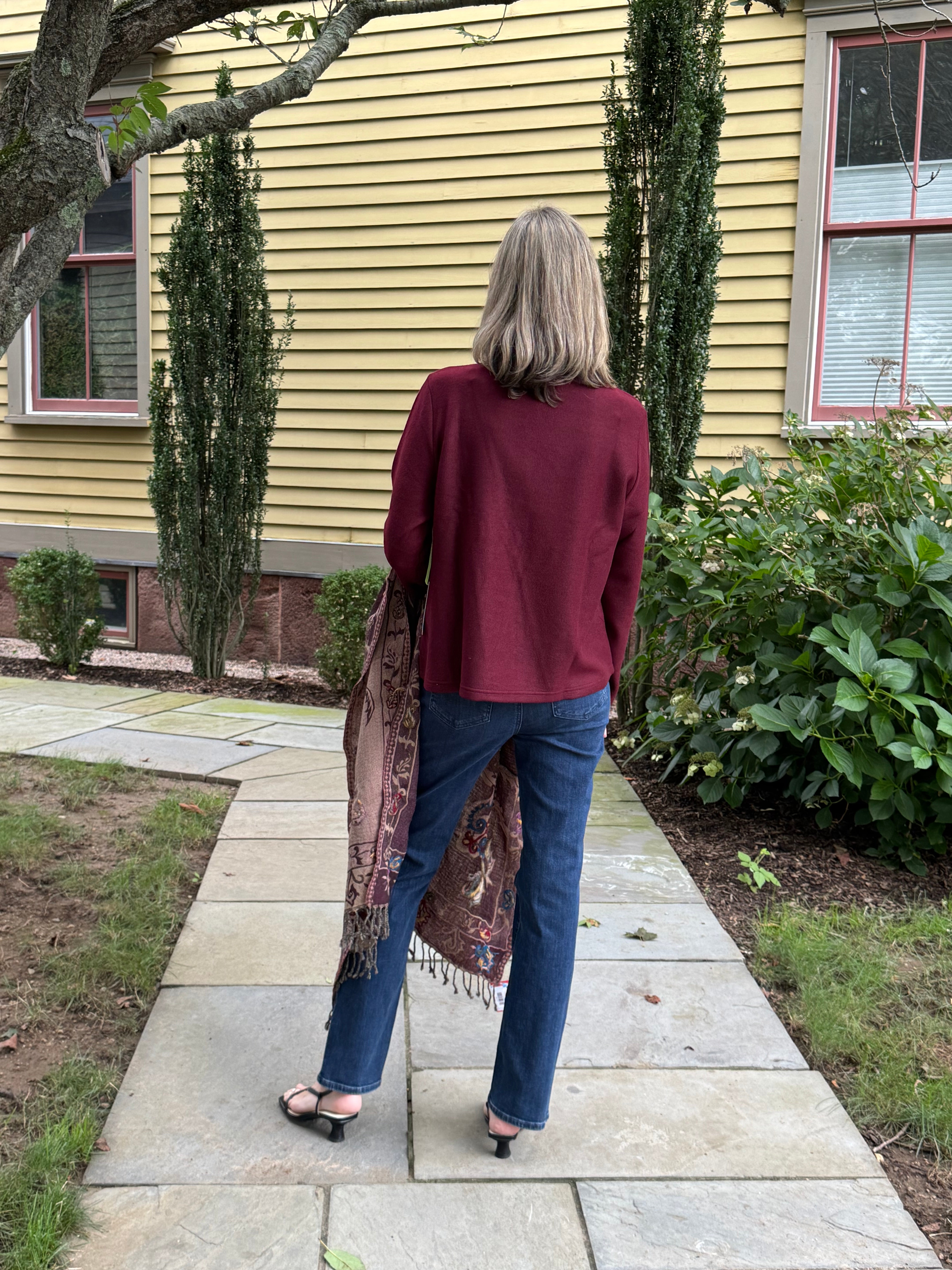 A woman viewed from behind stands on a stone walkway in front of a yellow house, wearing a burgundy long-sleeve top, blue jeans, and black sandals, while holding a patterned fringed scarf.