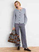 A woman modeling a light blue knit cardigan and gray trousers, holding a plaid tote bag against a plain white wall.
