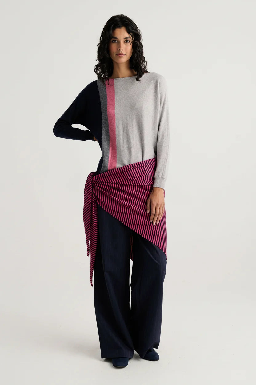 A model stands against a light gray studio background wearing a color-blocked knit sweater with navy, light gray, and pink sections, paired with dark wide-leg pants and a pink striped wrap tied around the waist.