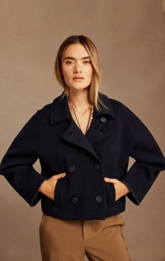 A woman modeling a cropped double-breasted navy wool-blend jacket with wide lapels and tan trousers, posed against a solid warm taupe background.