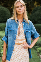 A woman standing outdoors in a garden wearing a light blue denim shirt over a cream patterned dress with a tan belt and a large white pendant necklace.