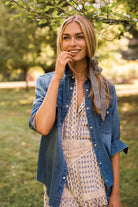 A woman standing outdoors in a park wearing a blue denim shirt over a beige patterned dress, with a gingham hair tie in her hair.