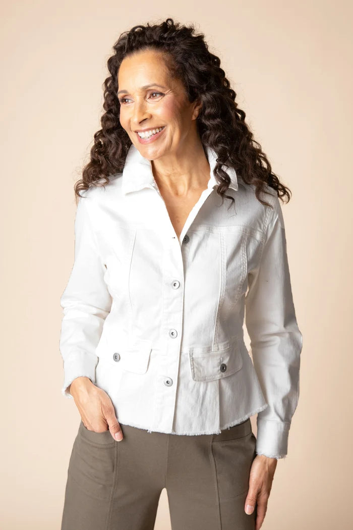 A smiling woman with long curly dark hair wearing a white button-front jacket and taupe pants, standing against a plain beige studio background.