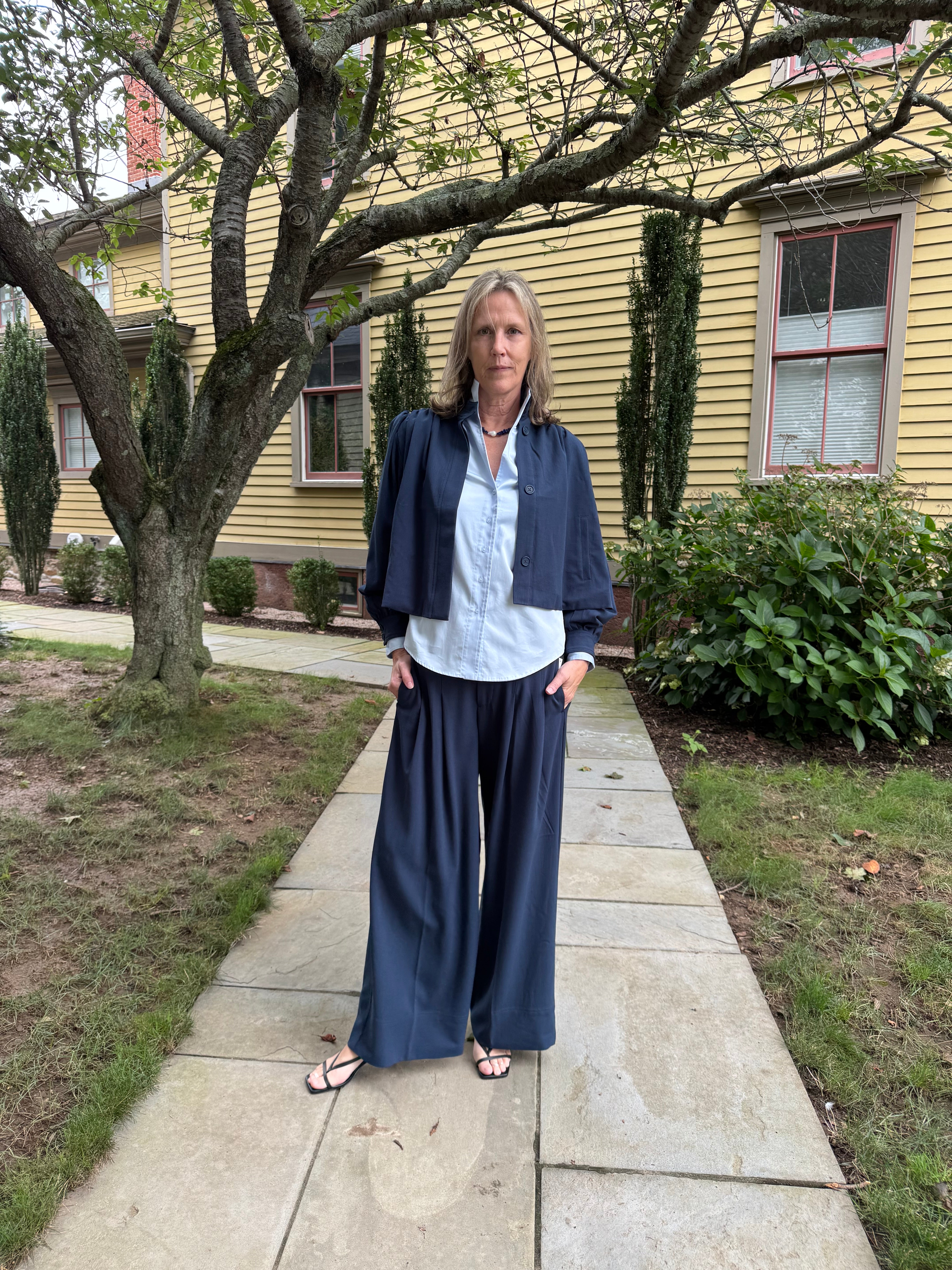A woman stands on a stone walkway in front of a yellow house, wearing a navy blue cropped jacket, light blue button-up shirt, and navy wide-leg pants.