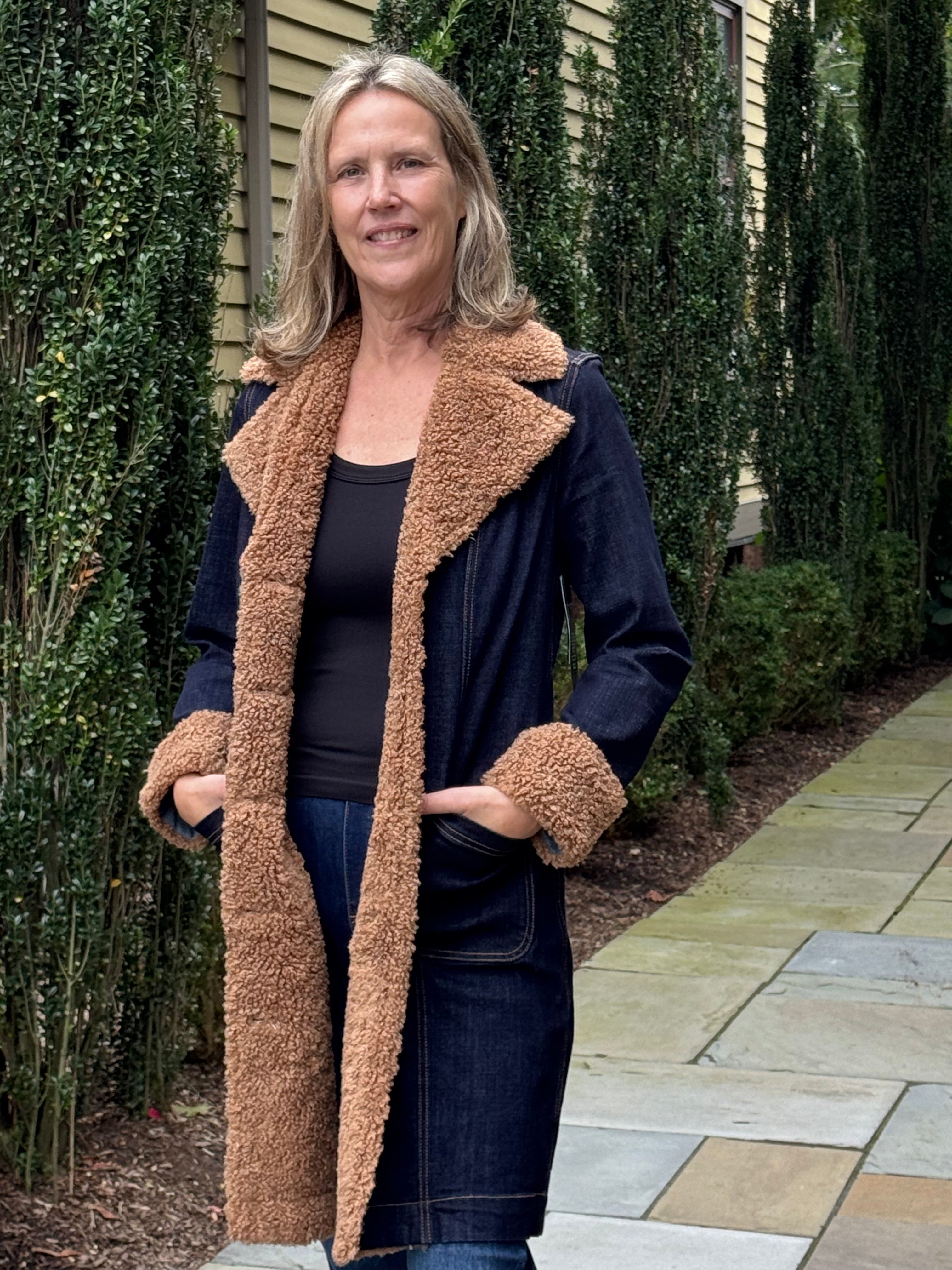 A woman wearing a dark blue denim-style coat with a thick tan sherpa lining, standing outdoors on a stone walkway with hedges and a house in the background.
