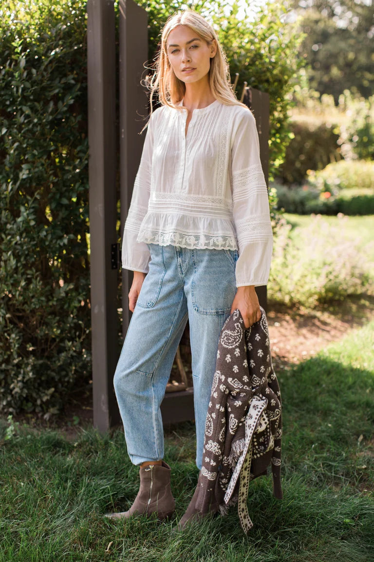 A woman stands outdoors on grass beside a wooden fence, wearing a white long-sleeve peplum blouse, light blue jeans, and brown ankle boots, while holding a dark paisley scarf.