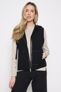 A woman modeling a black zip-up sleeveless vest over a light beige long-sleeve top and black pants, photographed against a plain light gray studio background.