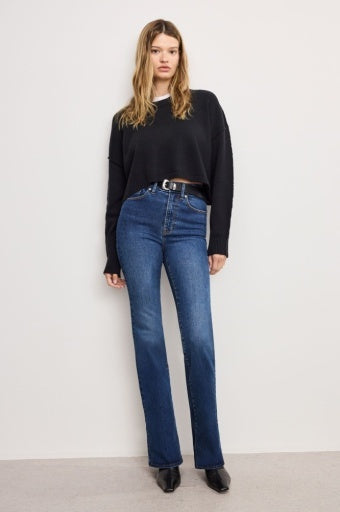 A woman modeling a black long-sleeve cropped sweater and high-waisted blue jeans with a black belt, standing against a plain off-white wall.