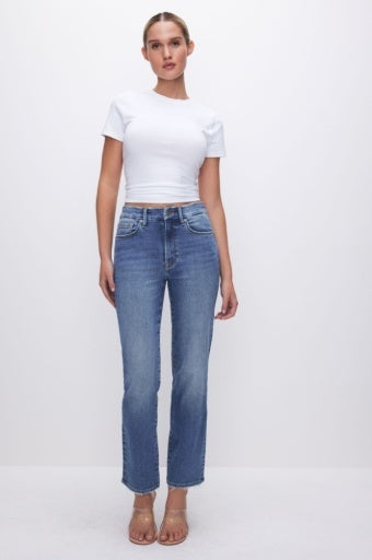 A woman modeling a pair of high-waisted, straight-leg blue jeans with a white cropped t-shirt and beige sandals, standing against a plain white studio background.