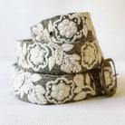 A woven belt with a gray and off-white floral pattern, coiled with a brass buckle on a light fabric background.