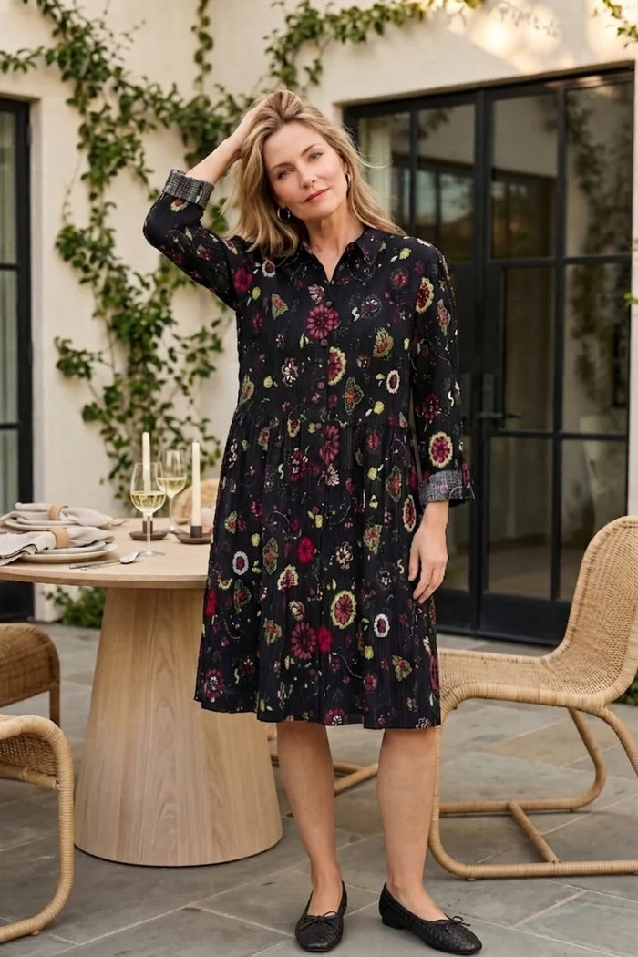 A woman stands on an outdoor patio wearing a dark floral midi dress with three-quarter sleeves and black flats. The background features a wooden table, wicker chairs, and a glass door with green ivy climbing on the wall.