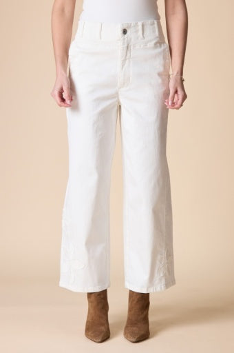 A model from the waist down wearing white cropped wide-leg jeans with tonal embroidery on the lower legs, paired with brown suede ankle boots against a solid beige background.