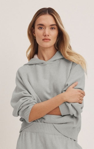 A woman wearing a light gray ribbed knit hoodie and matching sweatpants, posed against a solid off-white background.