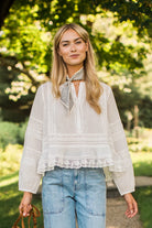 A woman wearing a white long-sleeve lace blouse with a patterned neck scarf, paired with light blue jeans, standing outdoors in a sunny park with green trees in the background.