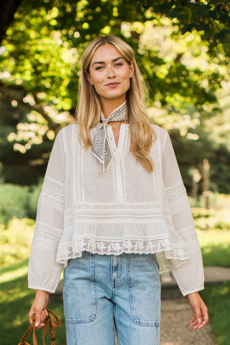 A woman wearing a white long-sleeve lace blouse with a patterned neck scarf, paired with light blue jeans, standing outdoors in a sunny park with green trees in the background.