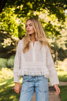 A woman wearing a white long-sleeve blouse with lace detailing and light blue jeans, standing outdoors in a sunlit park with green trees in the background.