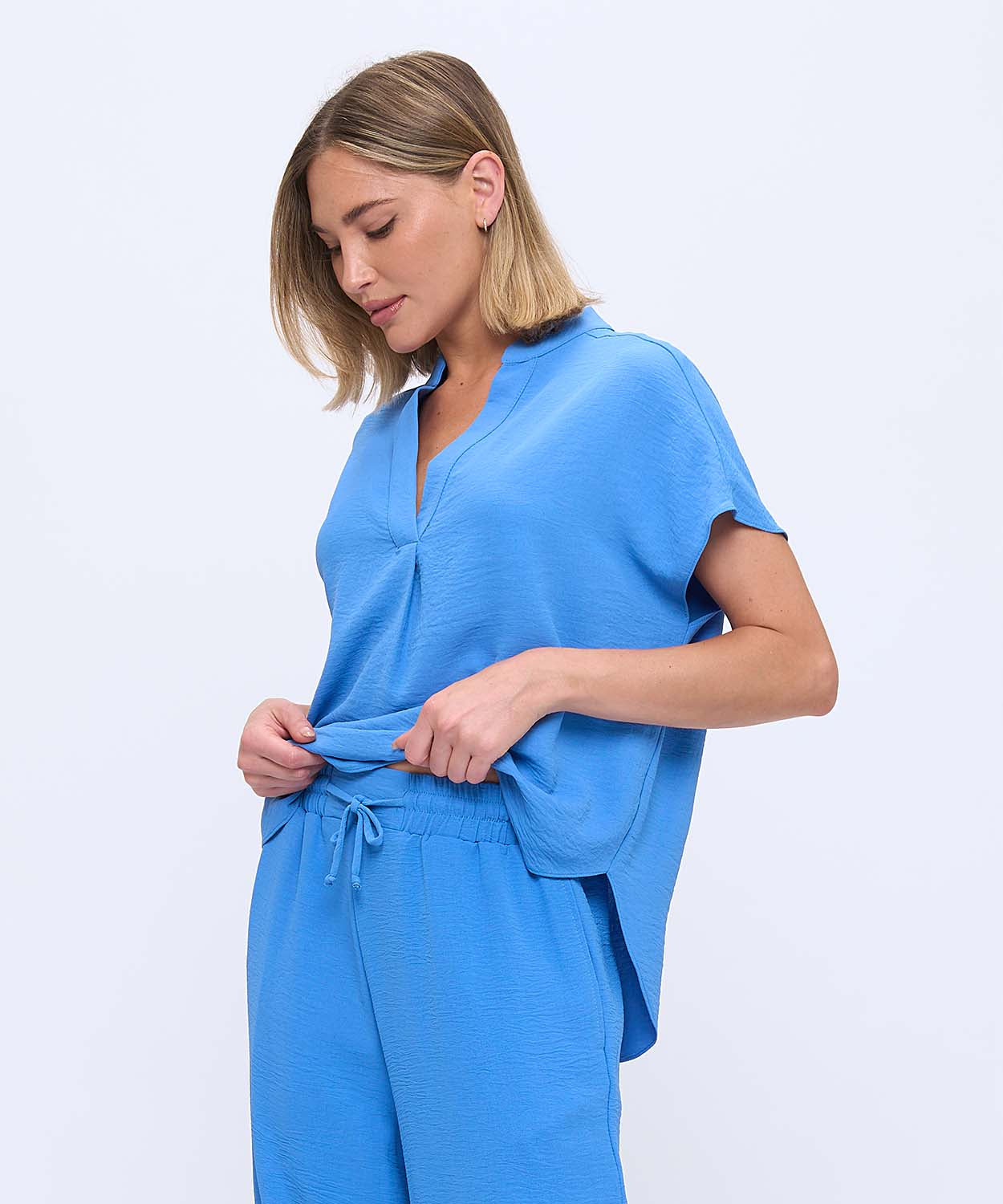 A woman wearing a bright blue short-sleeve top and matching drawstring pants, photographed against a clean light gray studio background.