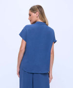 A woman shown from the back wearing a blue short-sleeve scrub top and matching blue pants against a light blue studio background.