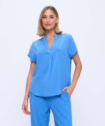 A woman modeling a bright blue scrub top and matching scrub pants against a plain light gray studio background.